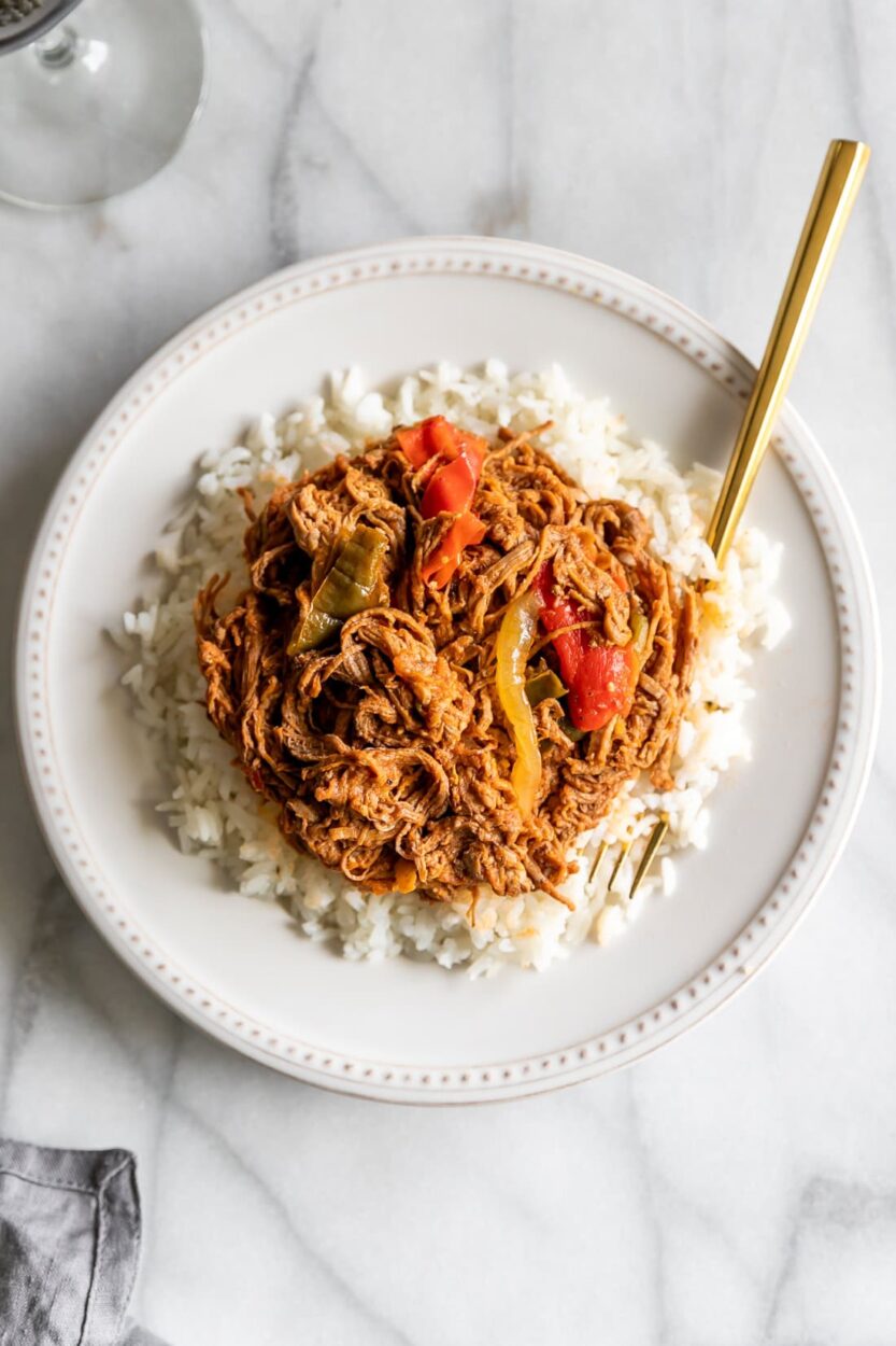 Cuban Ropa Vieja served over white rice in a rustic bowl, garnished with green olives and fresh cilantro, rich red tomato sauce glistening under warm light