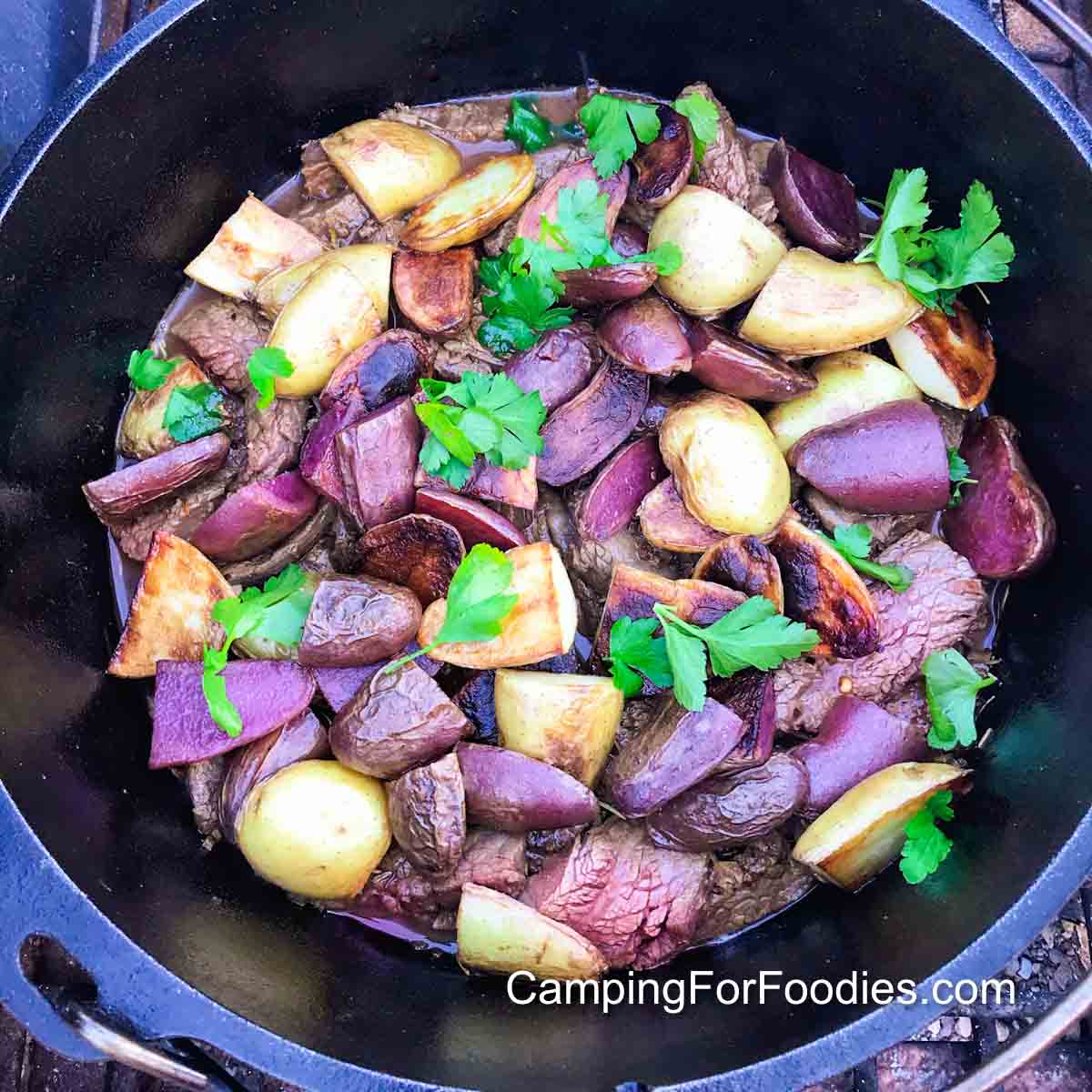 Raw flank steak being seared in a hot Dutch oven alongside bowls of prepped sofrito vegetables, spices, and green olives arranged on a wooden cutting board