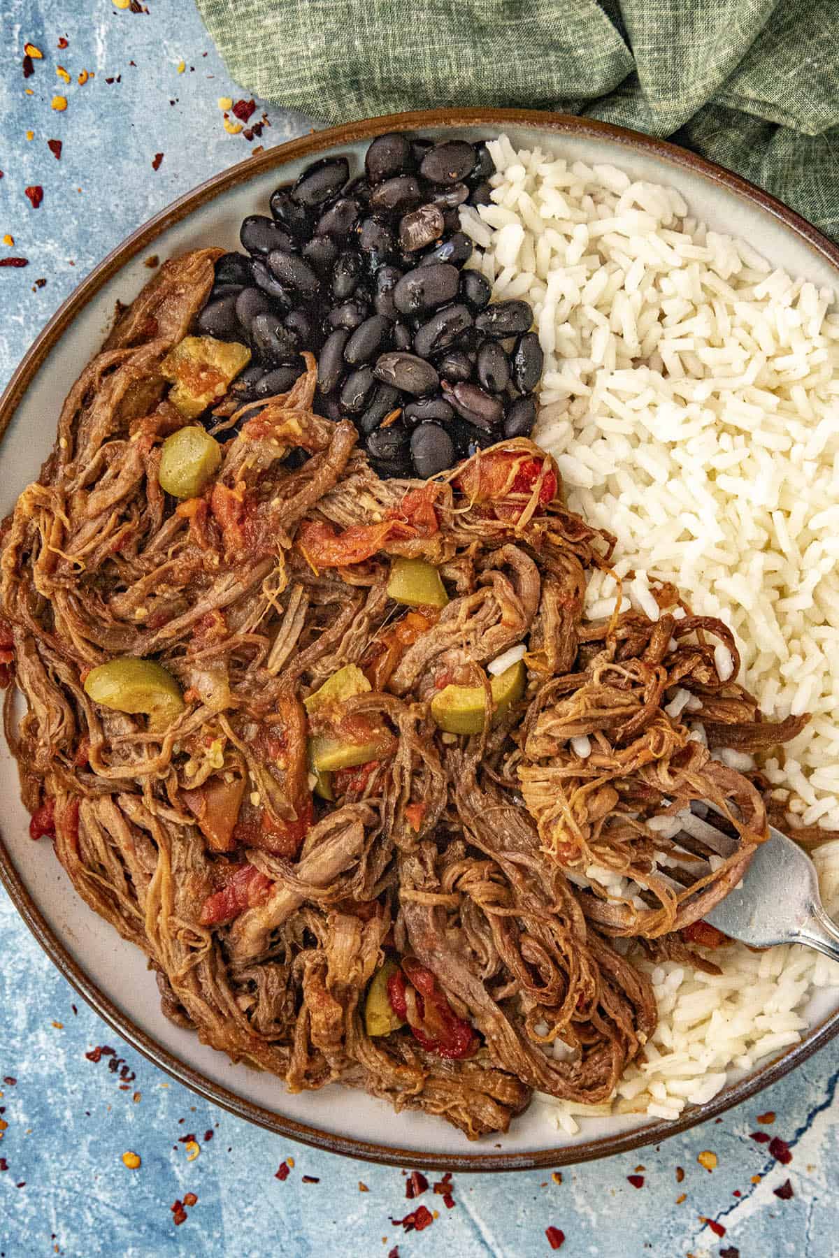 Close-up of Cuban Ropa Vieja — shredded braised beef pulled apart to show texture, nestled in glossy red tomato sauce with visible green olives, red pepper strips, and fresh herbs