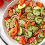 Cucumber tomato salad in a white bowl with red onion, fresh dill, and a glossy vinaigrette dressing — overhead shot on a rustic wooden table