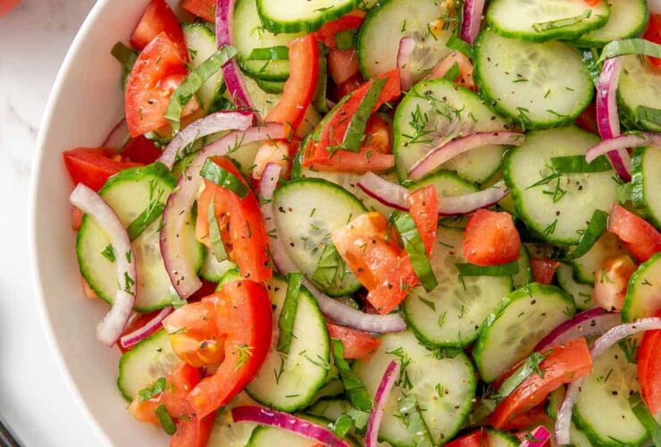 Cucumber tomato salad in a white bowl with red onion, fresh dill, and a glossy vinaigrette dressing — overhead shot on a rustic wooden table