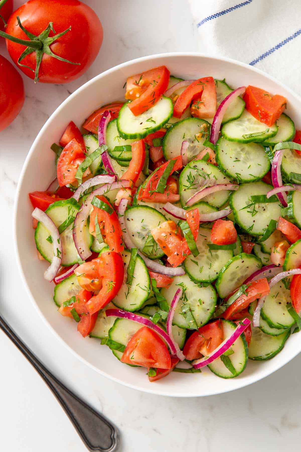 Cucumber tomato salad in a white bowl with red onion, fresh dill, and a glossy vinaigrette dressing — overhead shot on a rustic wooden table