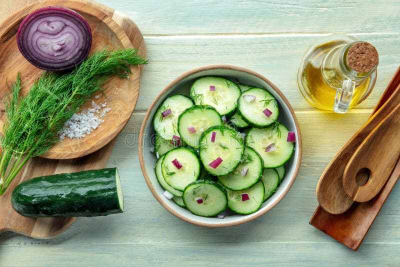 Sliced cucumbers, halved cherry tomatoes, thin-sliced red onion, fresh dill, and small bowls of olive oil and red wine vinegar arranged on a marble surface — ingredients flat lay