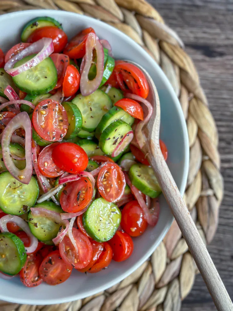 Close-up of cucumber tomato salad served in a small white bowl, glistening with vinaigrette, topped with fresh dill and cracked black pepper — shallow depth of field