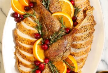 Whole dry brined roast turkey on a wooden carving board, golden brown shatteringly crisp skin glistening, garnished with fresh rosemary and thyme, dramatic side lighting