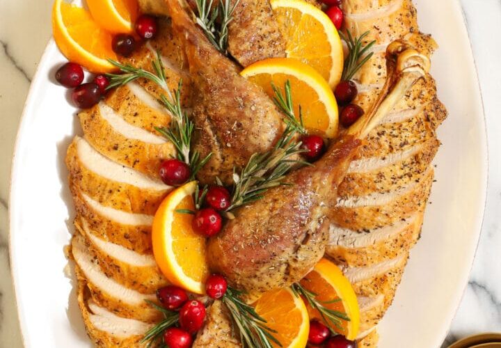 Whole dry brined roast turkey on a wooden carving board, golden brown shatteringly crisp skin glistening, garnished with fresh rosemary and thyme, dramatic side lighting