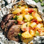 Foil packet steak and potato hash opened and steaming on a wooden board, showing golden potatoes, seared steak bites, and crispy bacon with fresh herb garnish