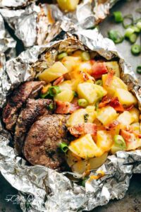 Foil packet steak and potato hash opened and steaming on a wooden board, showing golden potatoes, seared steak bites, and crispy bacon with fresh herb garnish