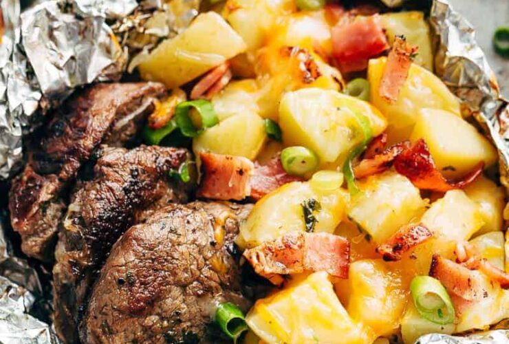 Foil packet steak and potato hash opened and steaming on a wooden board, showing golden potatoes, seared steak bites, and crispy bacon with fresh herb garnish