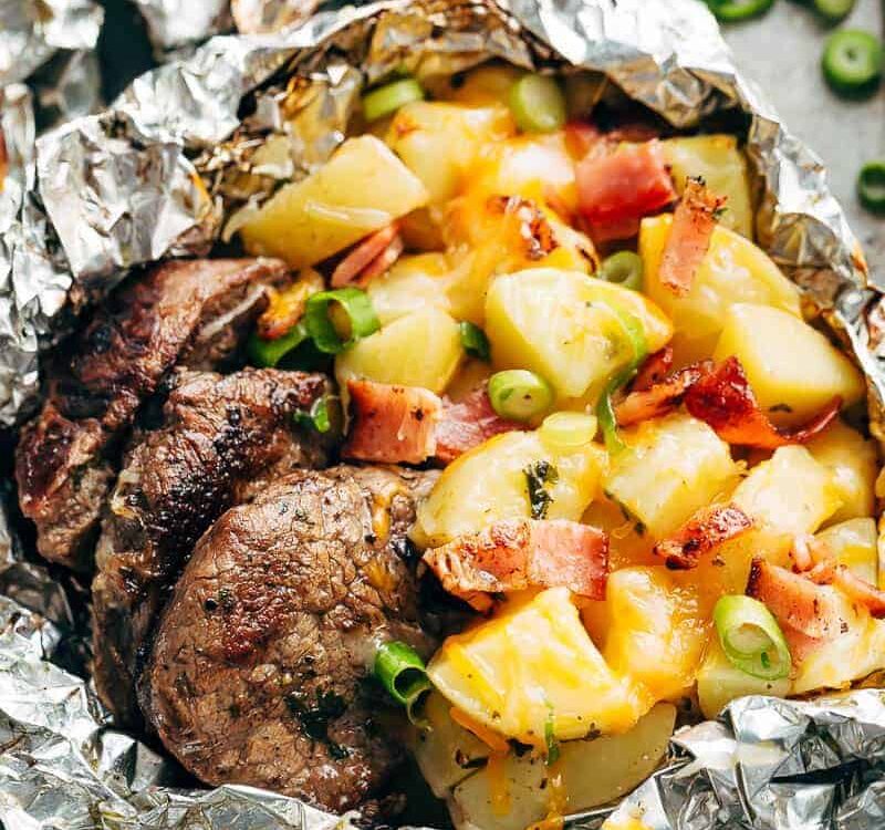 Foil packet steak and potato hash opened and steaming on a wooden board, showing golden potatoes, seared steak bites, and crispy bacon with fresh herb garnish