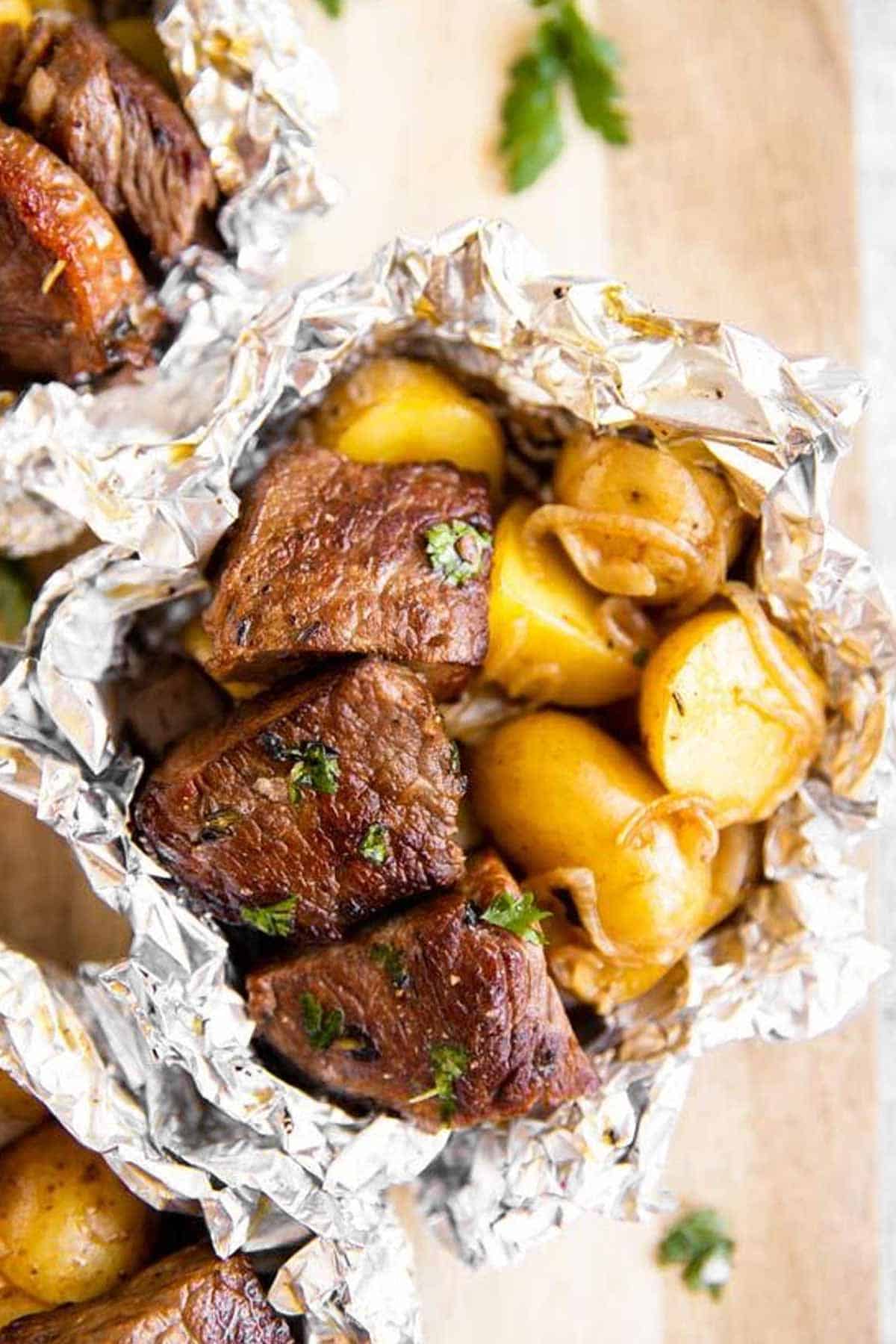 Overhead flat-lay of raw ingredients for foil packet steak and potato hash — diced sirloin, sliced potatoes, raw bacon strips, garlic, butter pats, and spices on a dark surface