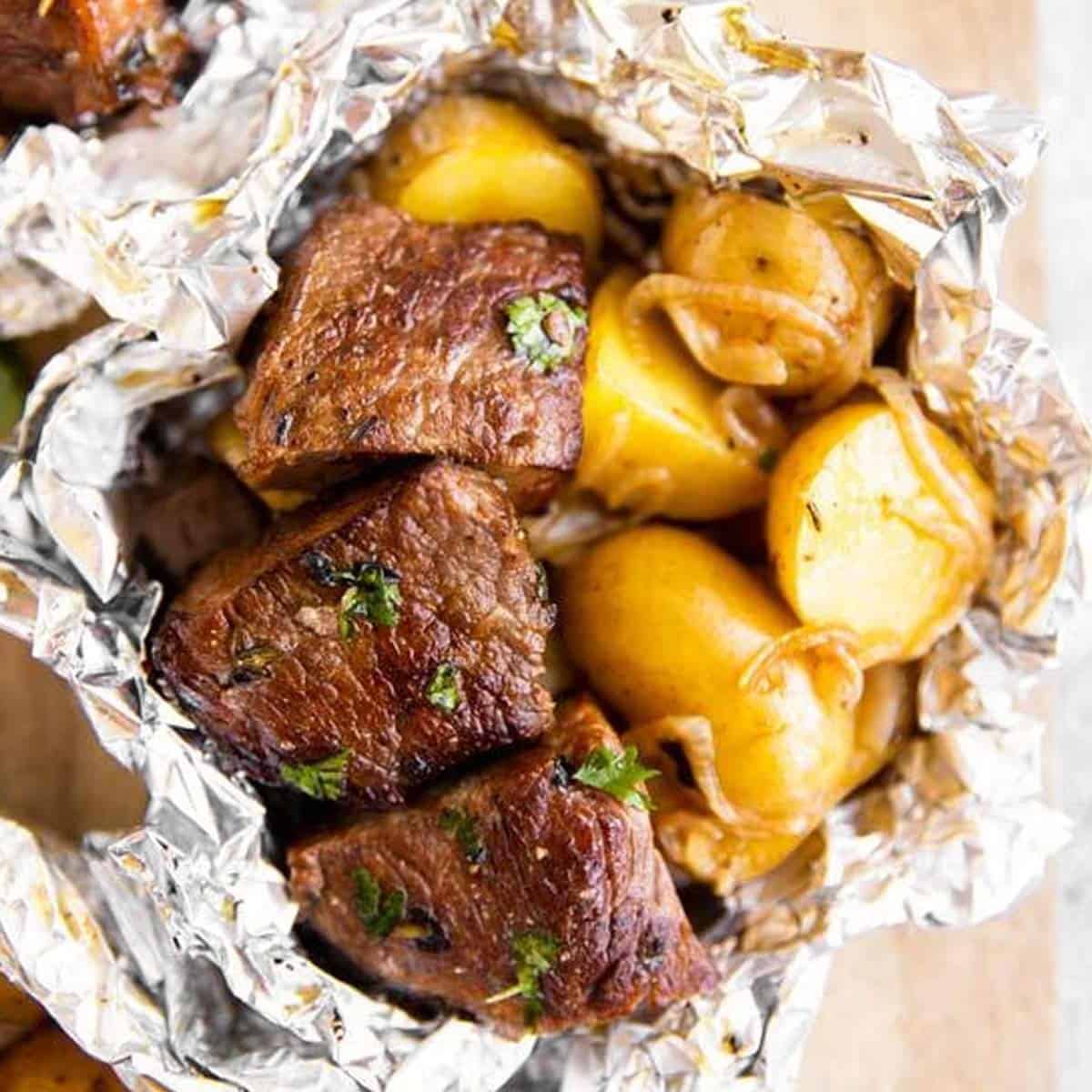Close-up of foil packet steak and potato hash served in the opened foil with a fork lifting a piece of steak, showing the glossy butter-bacon pan sauce and golden potato edges