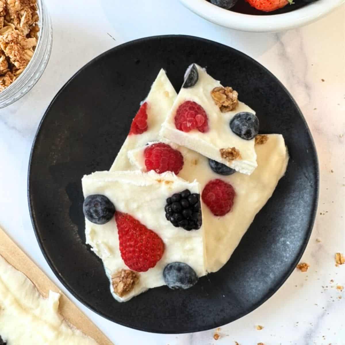 flat lay of ingredients for frozen greek yogurt bark including greek yogurt in a bowl, fresh mixed berries, honey jar, granola, and vanilla extract on a light wooden surface