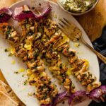 Greek Chicken Souvlaki skewers beautifully plated on a wooden board with pita bread, tzatziki, lemon wedges, and fresh herbs