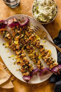 Greek Chicken Souvlaki skewers beautifully plated on a wooden board with pita bread, tzatziki, lemon wedges, and fresh herbs