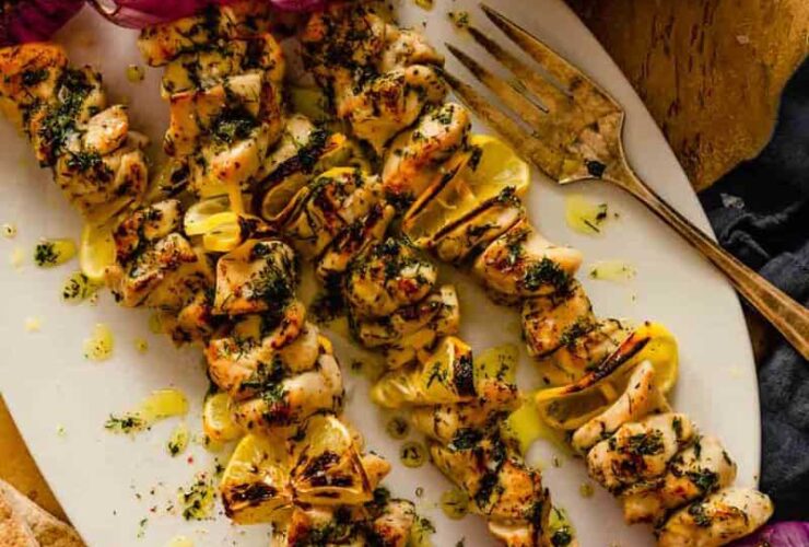 Greek Chicken Souvlaki skewers beautifully plated on a wooden board with pita bread, tzatziki, lemon wedges, and fresh herbs