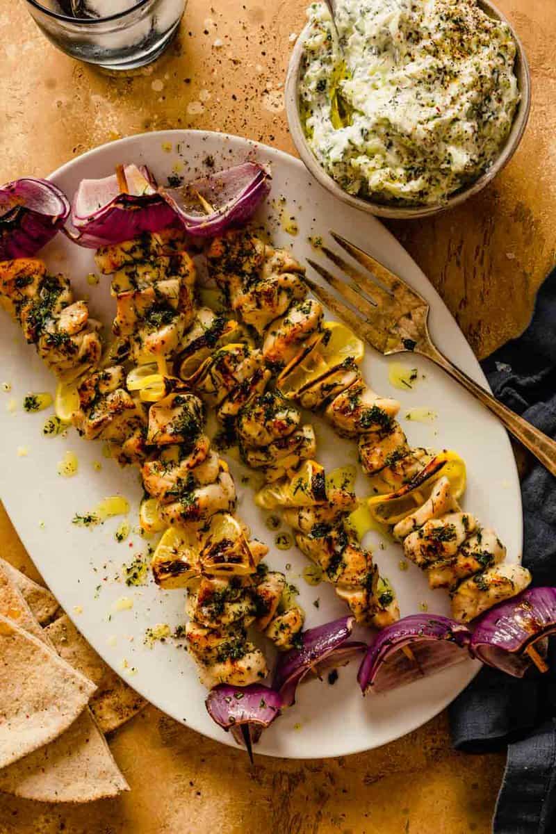 Greek Chicken Souvlaki skewers beautifully plated on a wooden board with pita bread, tzatziki, lemon wedges, and fresh herbs