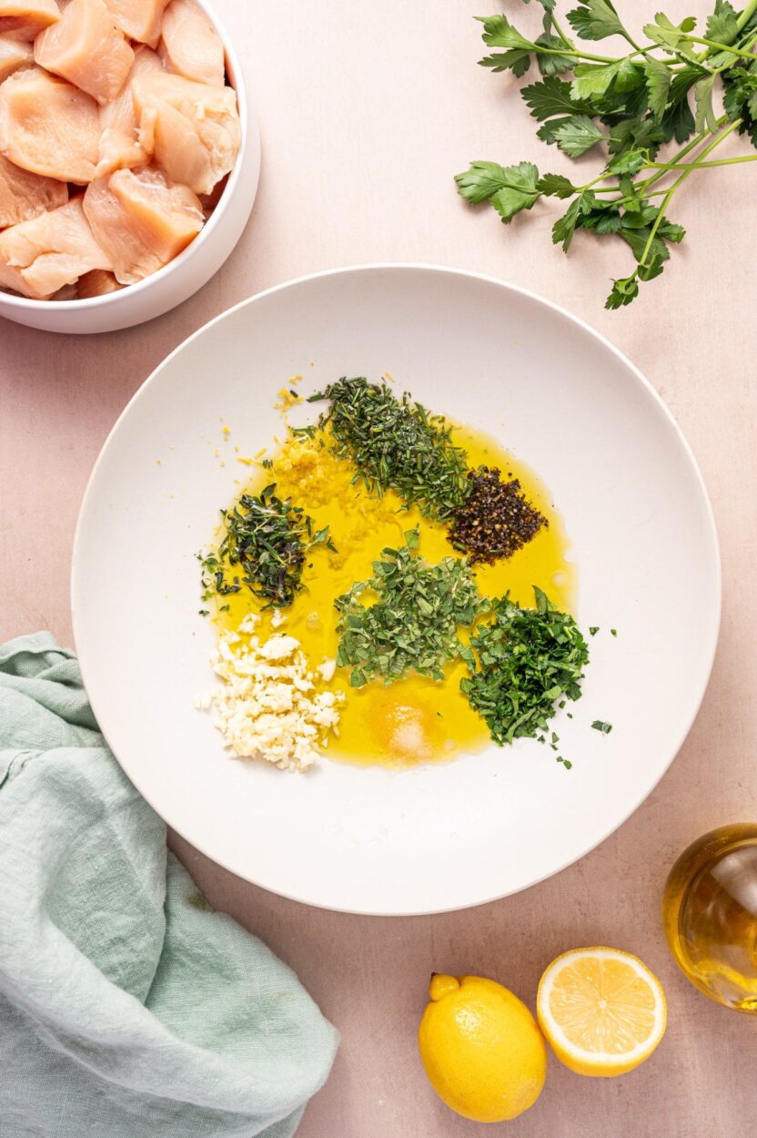 Greek Chicken Souvlaki marinade ingredients flat lay with chicken thighs, lemon, garlic, olive oil, oregano, and skewers on a marble surface
