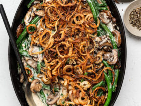 Green bean casserole from scratch in a white baking dish with golden crispy fried shallots on top, bubbling mushroom cream sauce visible at the edges, shot on a rustic wooden holiday table