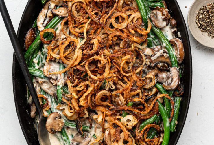 Green bean casserole from scratch in a white baking dish with golden crispy fried shallots on top, bubbling mushroom cream sauce visible at the edges, shot on a rustic wooden holiday table