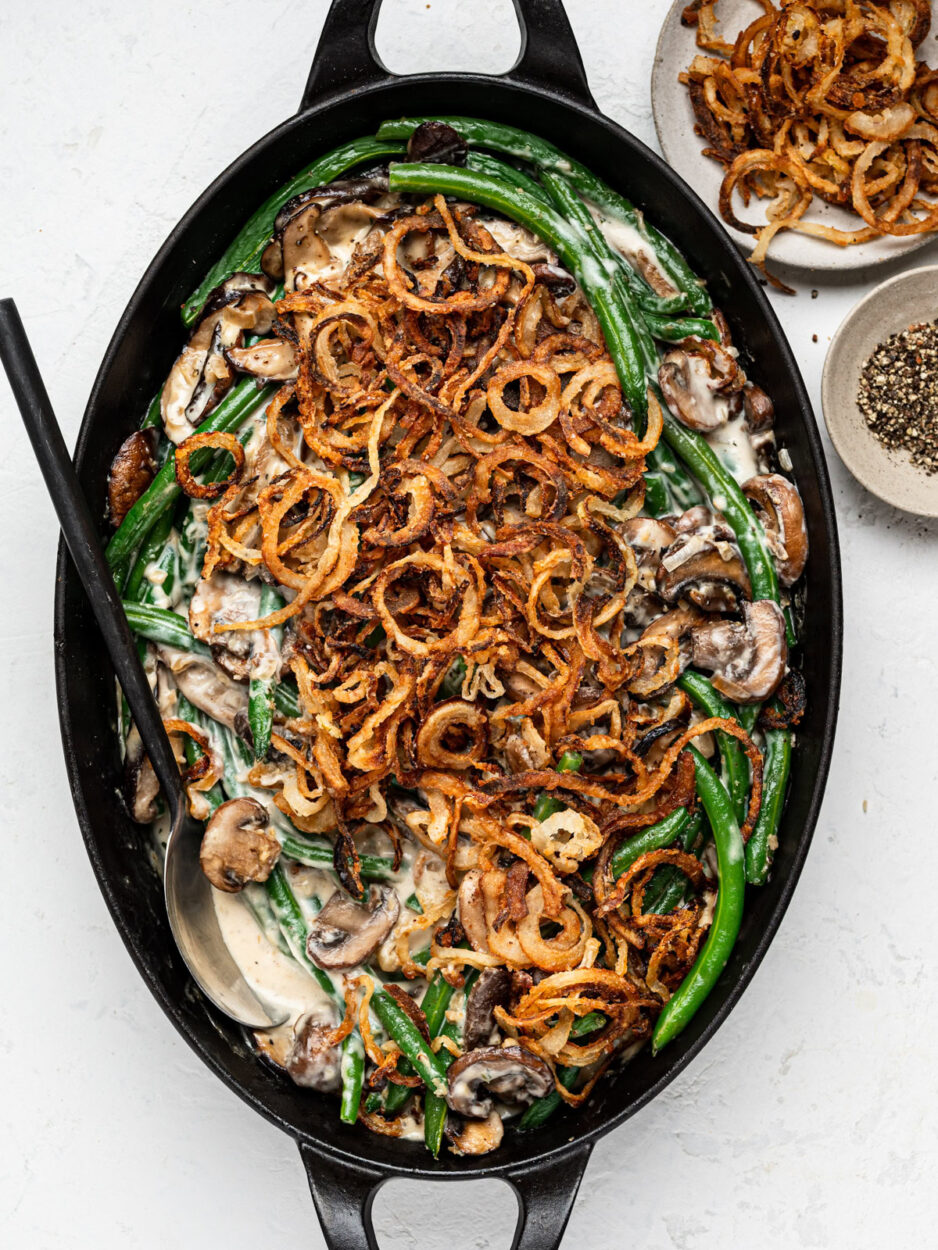 Green bean casserole from scratch in a white baking dish with golden crispy fried shallots on top, bubbling mushroom cream sauce visible at the edges, shot on a rustic wooden holiday table