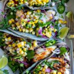 Grilled fish tacos with corn salsa served on a rustic wooden board, garnished with fresh cilantro, lime wedges, and charred corn