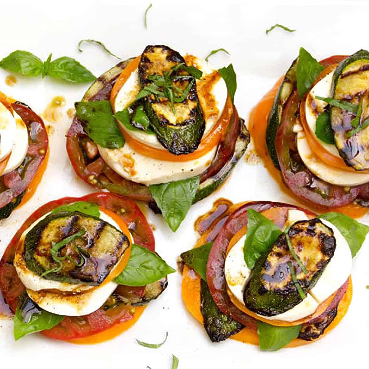 Overhead flat-lay of ingredients for grilled zucchini caprese stacks including sliced zucchini, fresh mozzarella ball, ripe tomatoes, basil, olive oil, and balsamic glaze on a wooden board