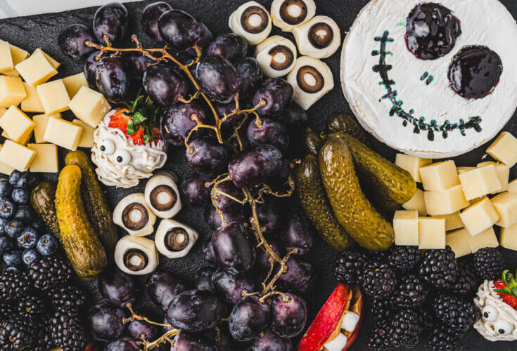 A beautifully styled Halloween charcuterie board with cheese ghosts, salami roses, black grapes, olives, and crackers arranged on a dark slate board with Halloween decorations
