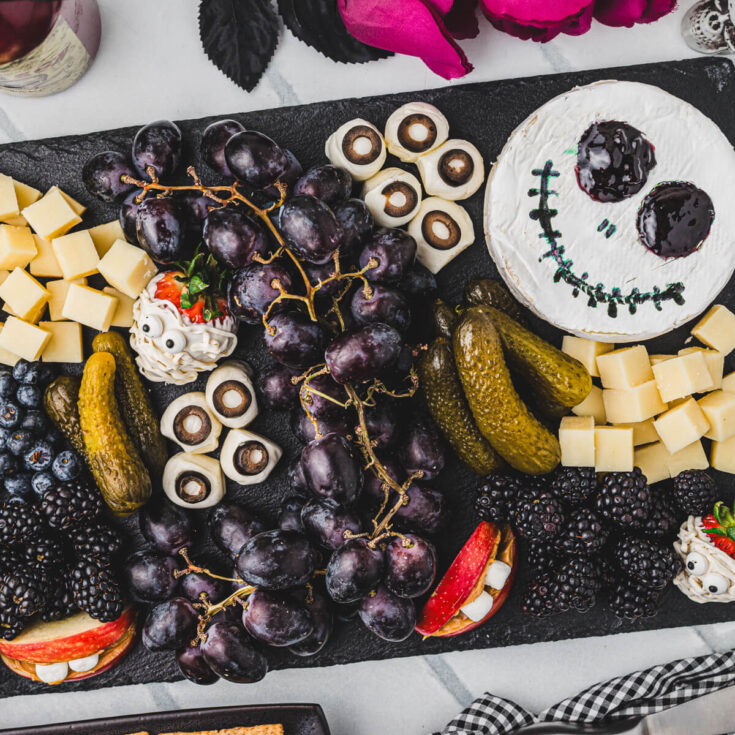 A beautifully styled Halloween charcuterie board with cheese ghosts, salami roses, black grapes, olives, and crackers arranged on a dark slate board with Halloween decorations