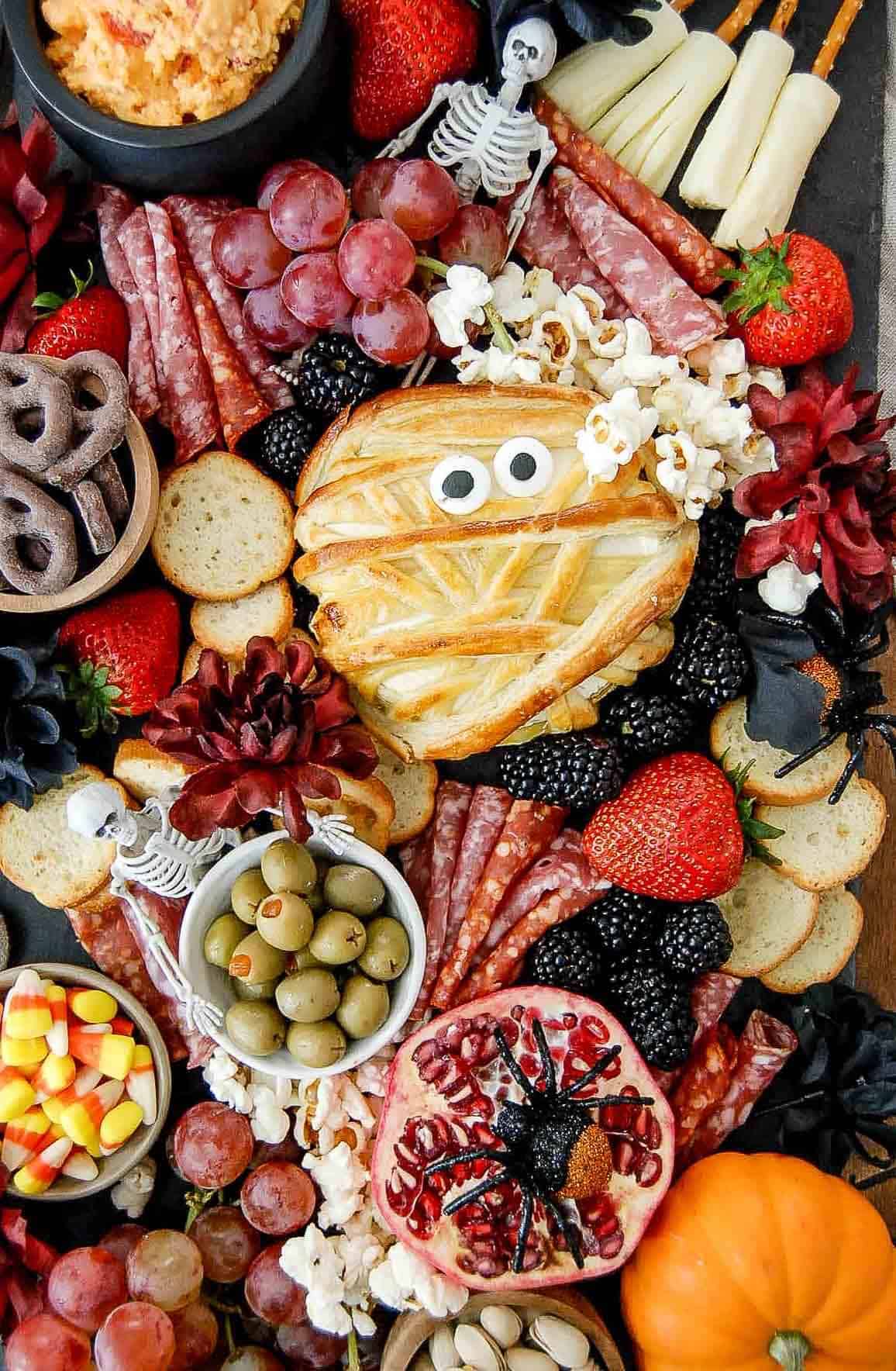Flat lay of Halloween charcuterie board ingredients including brie, salami, black grapes, cheddar, olives, and ghost-shaped cookie cutters on a dark surface