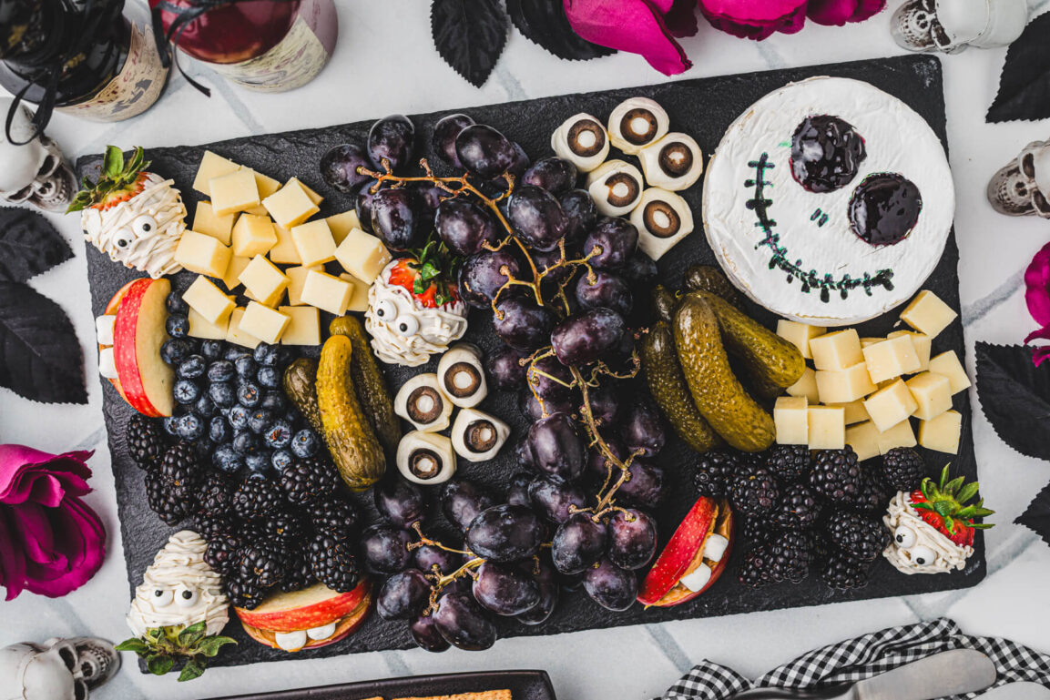 Close-up of cheese ghosts with candy eyes and salami roses on a Halloween charcuterie board, styled on a dark slate surface with orange and black accents