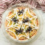 Halloween spider web taco dip in a clear dish with sour cream web design and olive spider on top, surrounded by tortilla chips