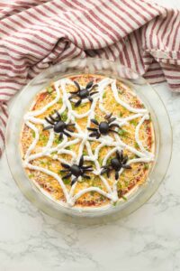 Halloween spider web taco dip in a clear dish with sour cream web design and olive spider on top, surrounded by tortilla chips