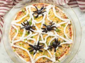 Halloween spider web taco dip in a clear dish with sour cream web design and olive spider on top, surrounded by tortilla chips