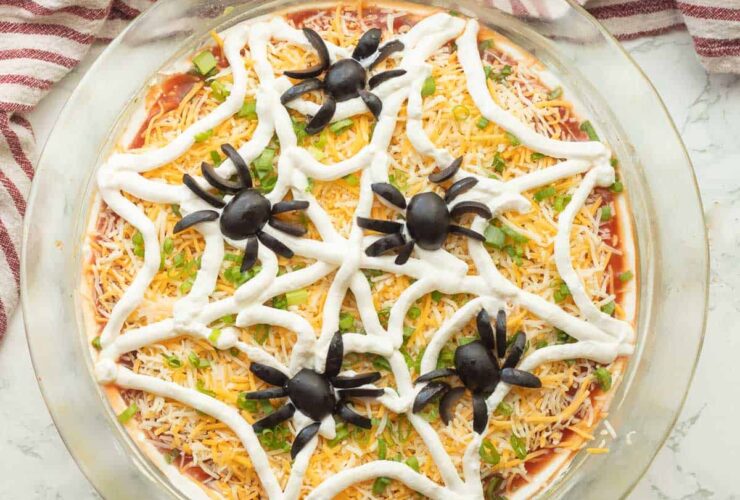 Halloween spider web taco dip in a clear dish with sour cream web design and olive spider on top, surrounded by tortilla chips