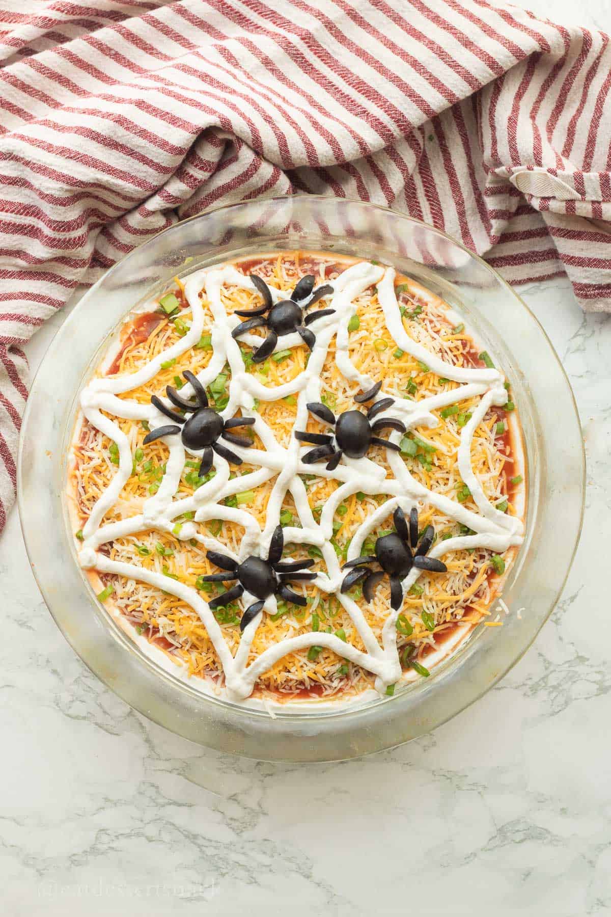 Halloween spider web taco dip in a clear dish with sour cream web design and olive spider on top, surrounded by tortilla chips