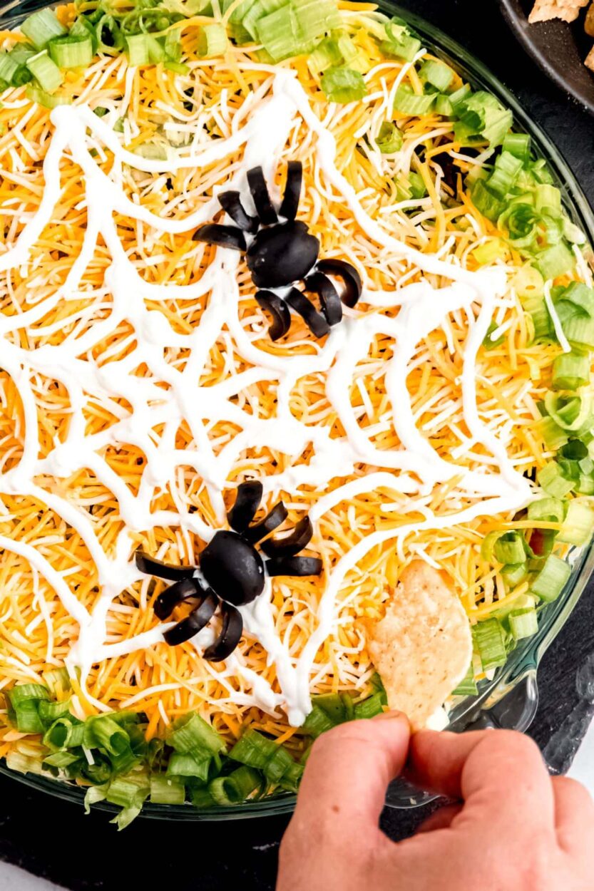 Close-up of a tortilla chip being pulled from the halloween spider web taco dip showing all seven layers — beans, beef, guacamole, salsa, cheese, and sour cream
