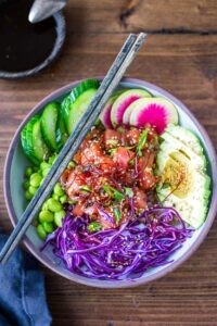 Hawaiian Poke Bowl beautifully plated in a white bowl with glossy soy-marinated ahi tuna, avocado slices, edamame, cucumber, pickled ginger, sriracha mayo drizzle, and sesame seeds on sushi rice