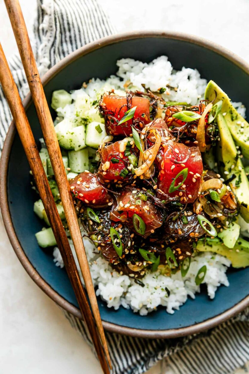 Overhead flat-lay of Hawaiian Poke Bowl ingredients including diced ahi tuna, avocado, cucumber, edamame, pickled ginger, soy sauce, sesame oil, rice, and toppings arranged on a marble surface