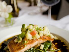 Herb-crusted salmon fillet with golden herb crust resting on a bed of roasted summer vegetables including zucchini, cherry tomatoes, and bell peppers, plated on a white dish with lemon wedges and fresh parsley
