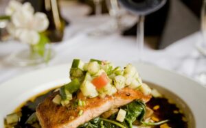 Herb-crusted salmon fillet with golden herb crust resting on a bed of roasted summer vegetables including zucchini, cherry tomatoes, and bell peppers, plated on a white dish with lemon wedges and fresh parsley