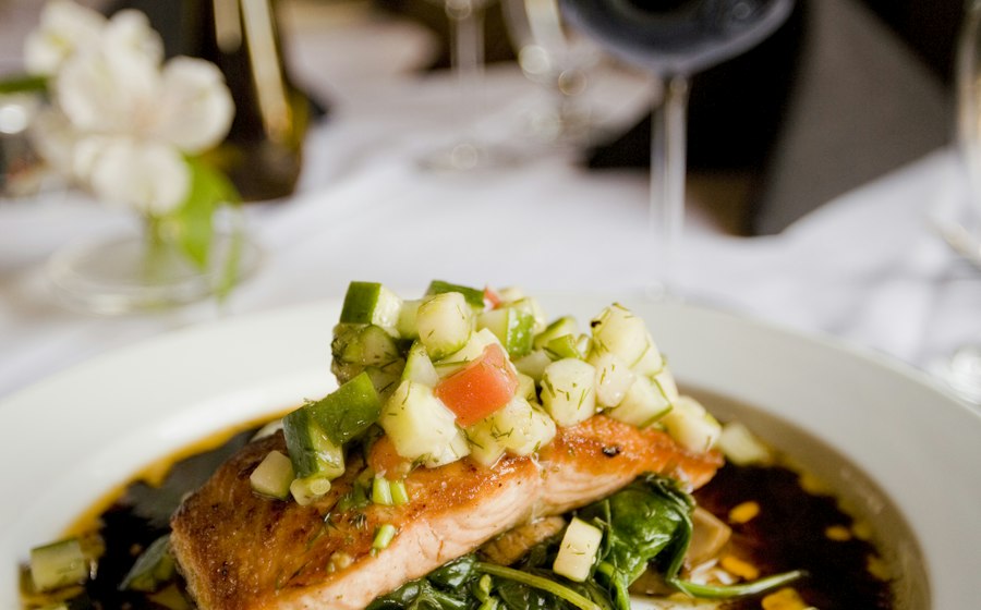 Herb-crusted salmon fillet with golden herb crust resting on a bed of roasted summer vegetables including zucchini, cherry tomatoes, and bell peppers, plated on a white dish with lemon wedges and fresh parsley