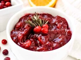 Homemade cranberry sauce in a white ceramic bowl with orange zest garnish and fresh rosemary on a rustic Thanksgiving table