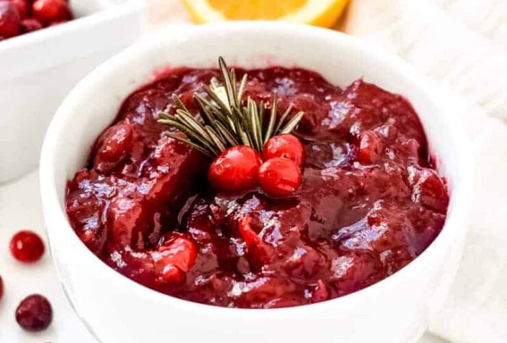 Homemade cranberry sauce in a white ceramic bowl with orange zest garnish and fresh rosemary on a rustic Thanksgiving table