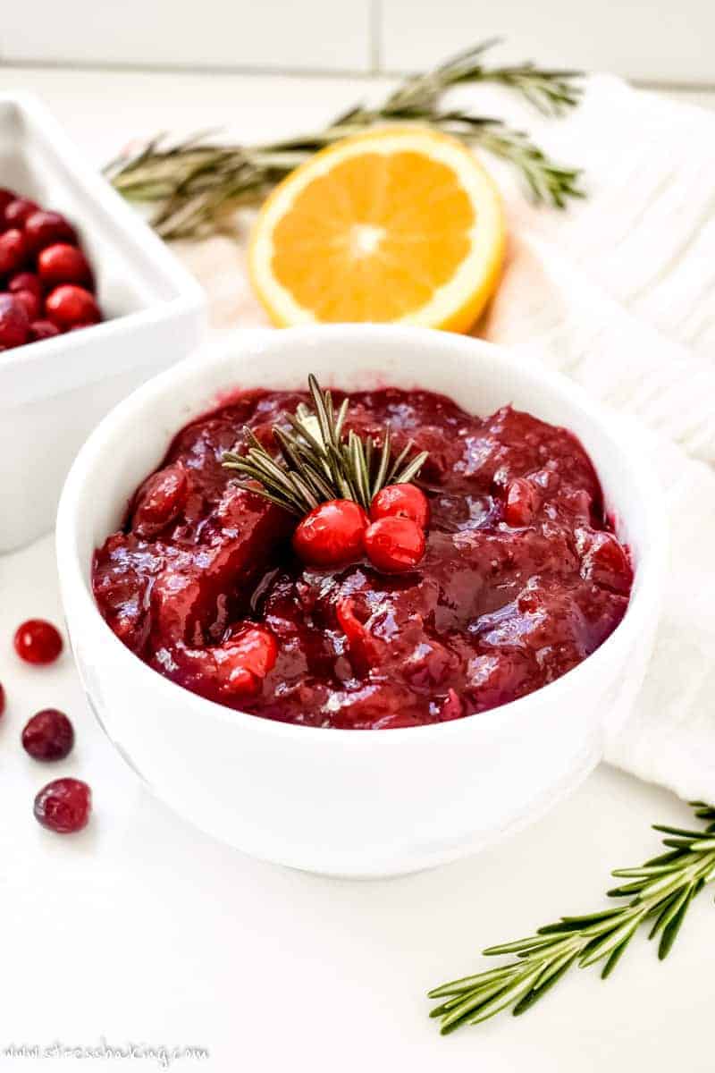 Homemade cranberry sauce in a white ceramic bowl with orange zest garnish and fresh rosemary on a rustic Thanksgiving table