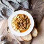 A deep bowl of Hungarian Goulash with fall-apart beef chunks in a rich paprika-red sauce, garnished with a dollop of sour cream and fresh parsley, served over egg noodles