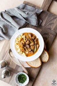 A deep bowl of Hungarian Goulash with fall-apart beef chunks in a rich paprika-red sauce, garnished with a dollop of sour cream and fresh parsley, served over egg noodles