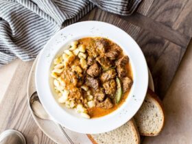 A deep bowl of Hungarian Goulash with fall-apart beef chunks in a rich paprika-red sauce, garnished with a dollop of sour cream and fresh parsley, served over egg noodles