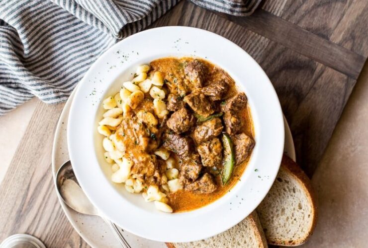 A deep bowl of Hungarian Goulash with fall-apart beef chunks in a rich paprika-red sauce, garnished with a dollop of sour cream and fresh parsley, served over egg noodles