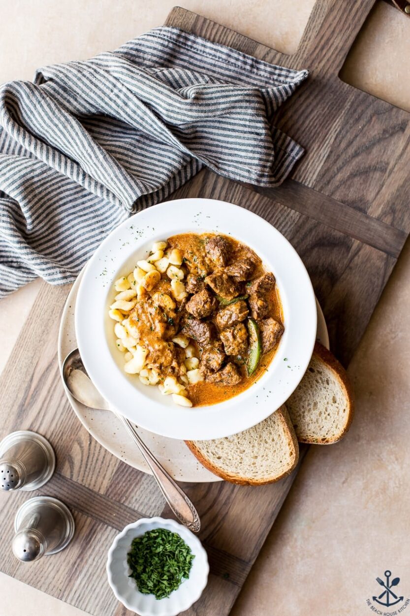 A deep bowl of Hungarian Goulash with fall-apart beef chunks in a rich paprika-red sauce, garnished with a dollop of sour cream and fresh parsley, served over egg noodles